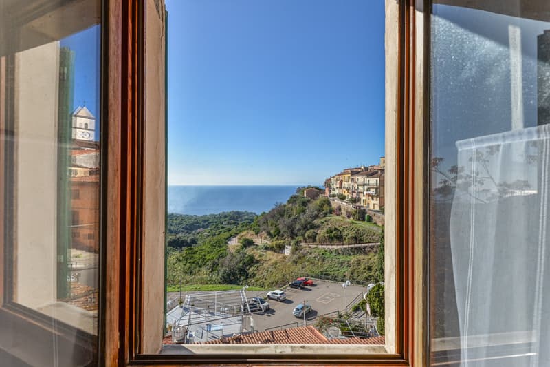 The image shows a scenic view of a coastal town from a window. The town is built on a hillside overlooking the ocean, with colorful buildings and lush greenery. A parking lot is visible in the foreground, and the clear blue sky and distant horizon can be