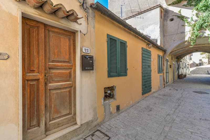 The image shows a narrow street in an old European town. The buildings have a traditional Mediterranean style, with yellow walls, green shutters, and a tiled roof. The street is paved with cobblestones, and there is a wooden door with an intercom system v