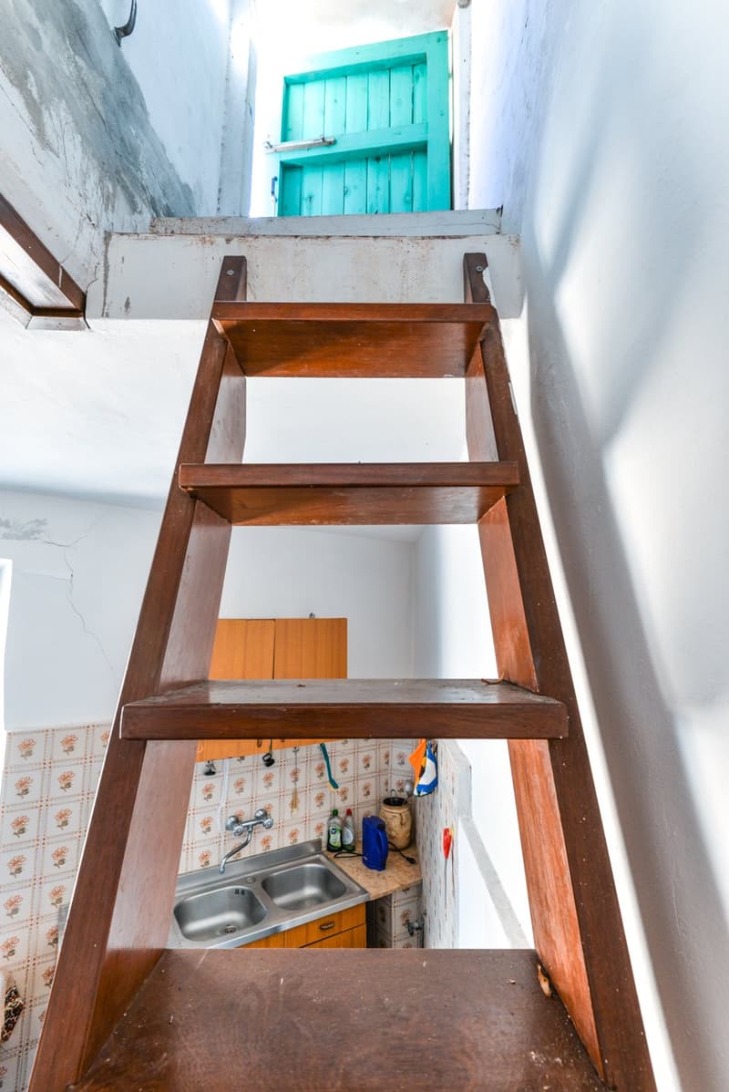 Wooden ladder leading to a small kitchenette area with a sink, storage shelves, and a tiled backsplash. The kitchenette is located in a narrow space with a turquoise wooden door visible at the top.