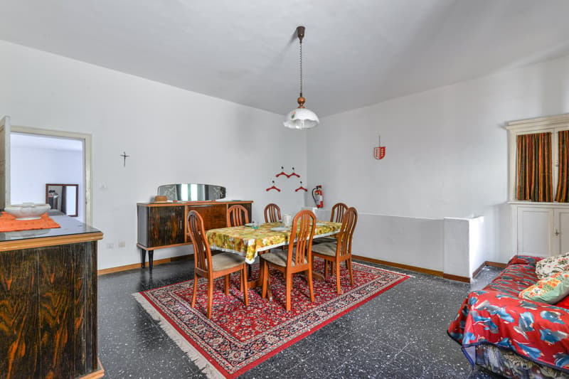 The image shows a dining room with a wooden dining table and chairs, a sideboard, and a patterned rug on the floor. The walls are painted in a light color, and there is a hanging light fixture above the dining table.