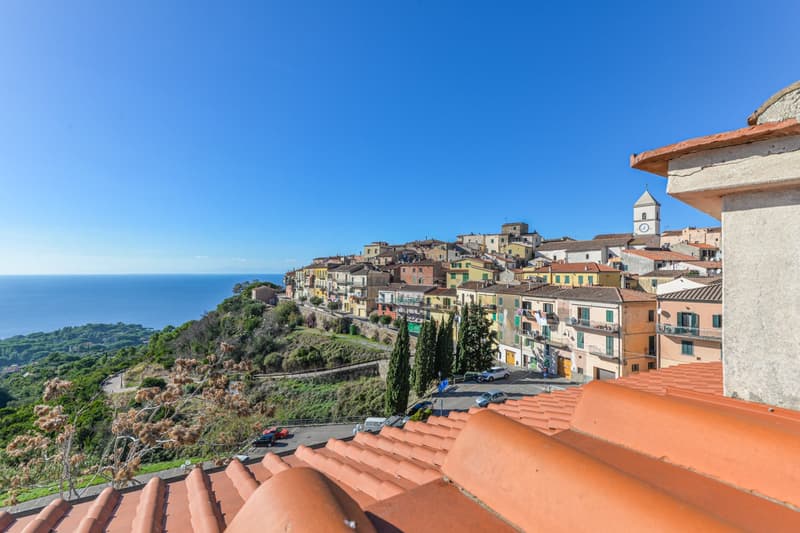 The image shows a picturesque coastal town built on a hillside, with colorful buildings, red-tiled roofs, and a church steeple visible. The town is surrounded by lush greenery and overlooks the sparkling blue waters of the sea in the distance.