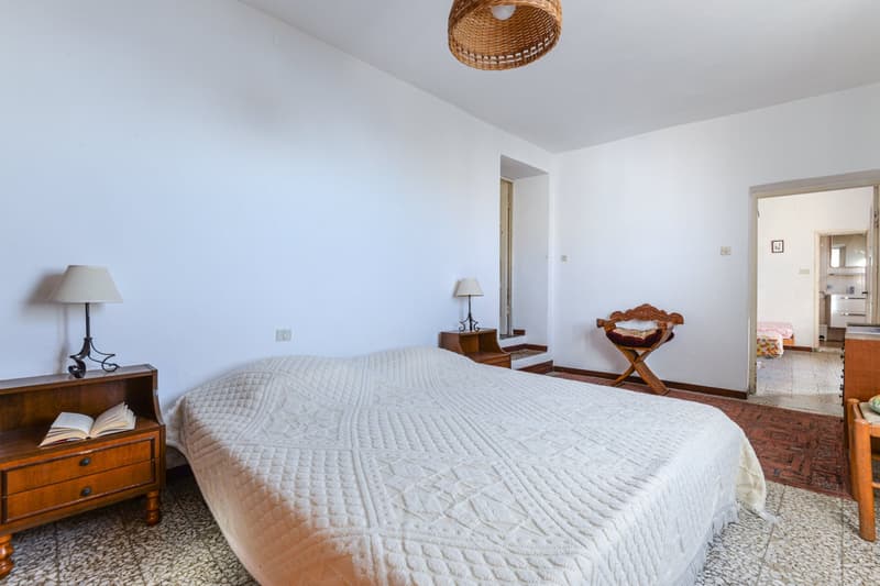 Spacious bedroom with white walls, hardwood floors, a large bed with a quilted bedspread, a wooden nightstand with a lamp, and a wicker pendant light fixture. There is also a wooden bench at the foot of the bed.