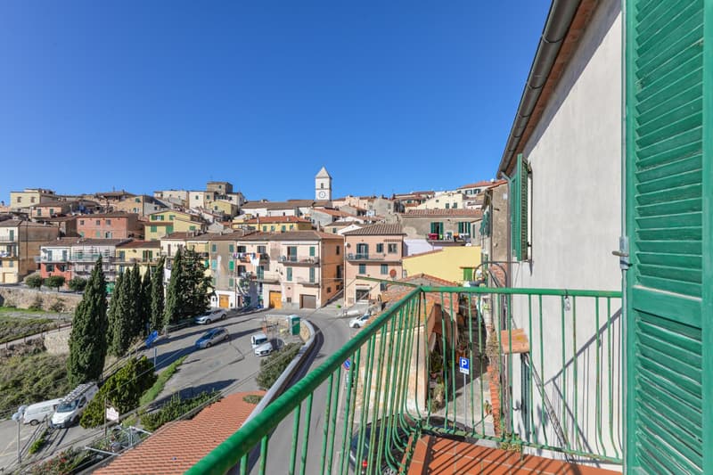 The image shows a picturesque Italian town with colorful buildings, narrow streets, and a church tower in the distance. The view is from a balcony or terrace, providing a panoramic perspective of the town nestled among the hills.