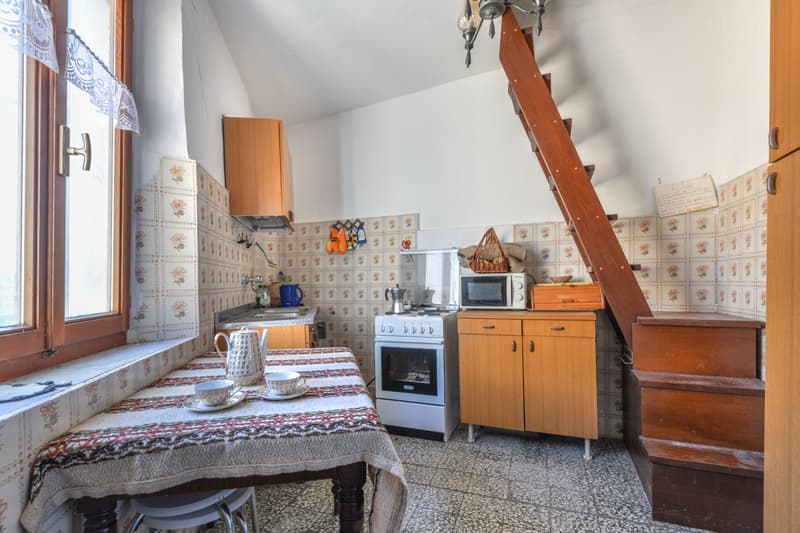 Tiled kitchen with wooden cabinets, stove, microwave, and dining table with patterned tablecloth. Wooden ladder leading to upper level.