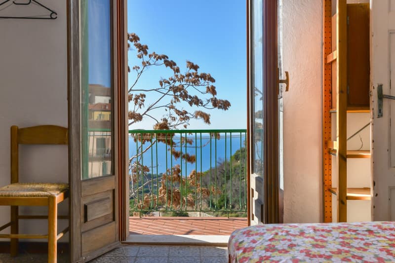 Bedroom with large windows overlooking a balcony or terrace, offering a scenic view of the surrounding landscape and water body. The room has wooden furniture and a floral-patterned bedding.