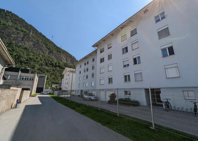 Multi-story apartment building with white exterior, surrounded by mountains and greenery, with a parking area in the foreground