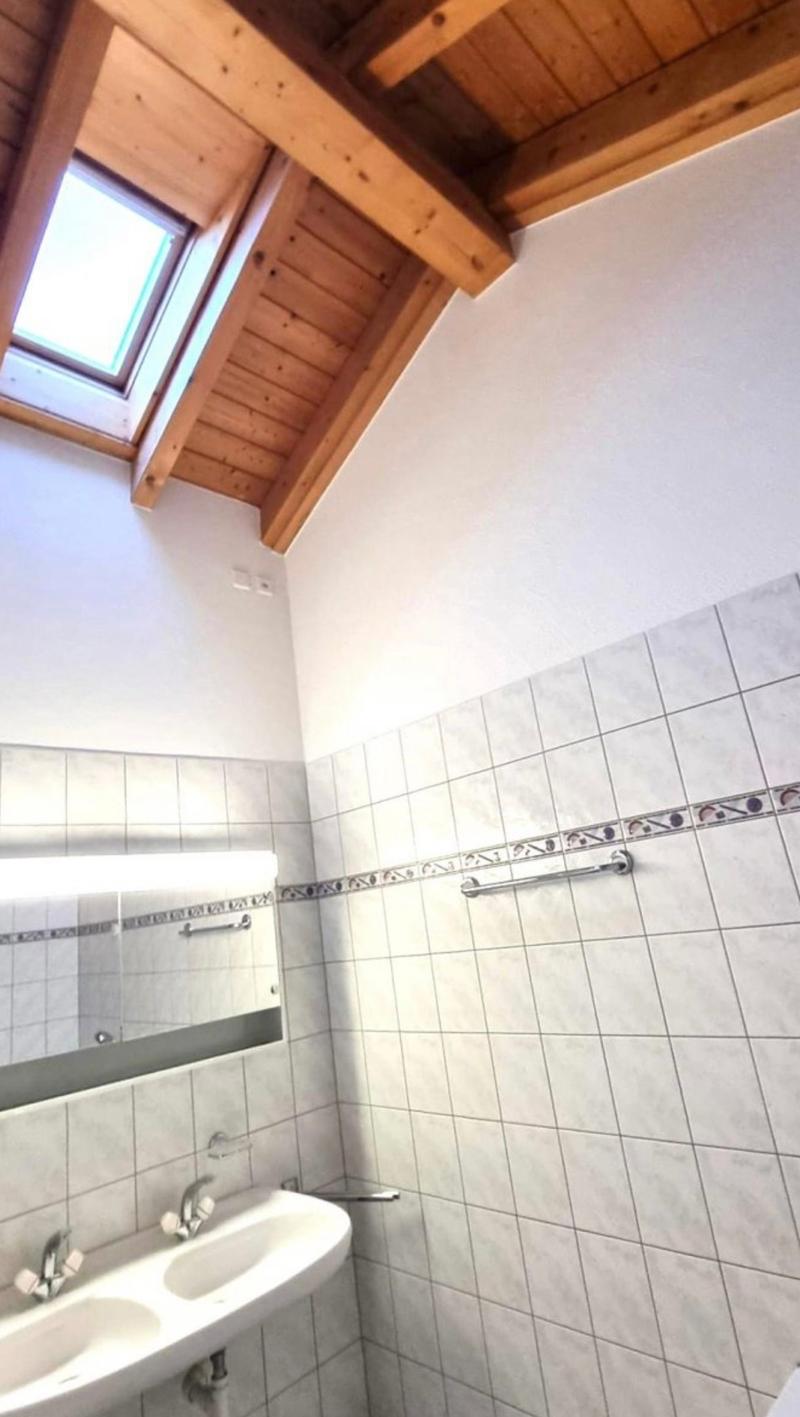 The image shows a bathroom with a tiled floor and walls, a sink, and a window with a wooden frame in the ceiling. The bathroom has a rustic, cabin-like feel with the exposed wooden beams and ceiling.