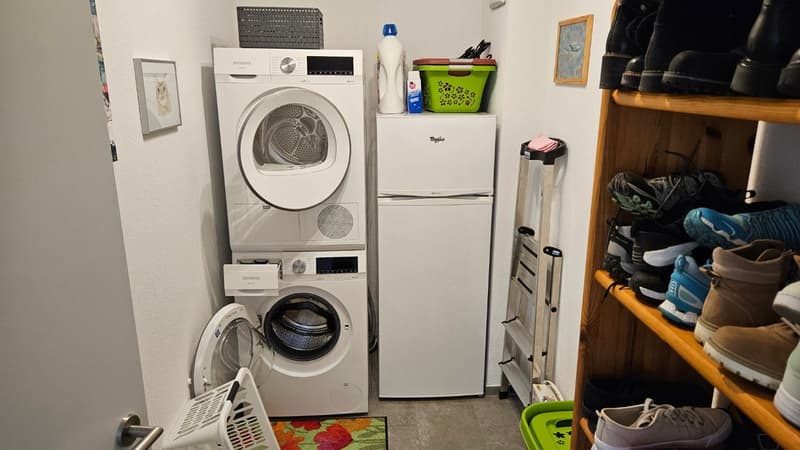 A small room with a washing machine, dryer, refrigerator, and a wooden shelf with shoes.