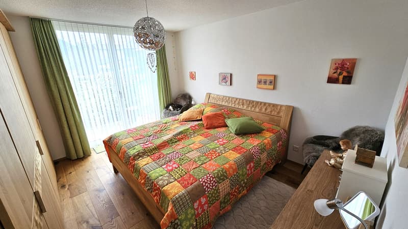 Bedroom with bed, wooden headboard, colorful quilt, pillows, rug, wooden nightstand, lamp, wall art, large window with curtains, wooden cabinet, and a view of the outside.