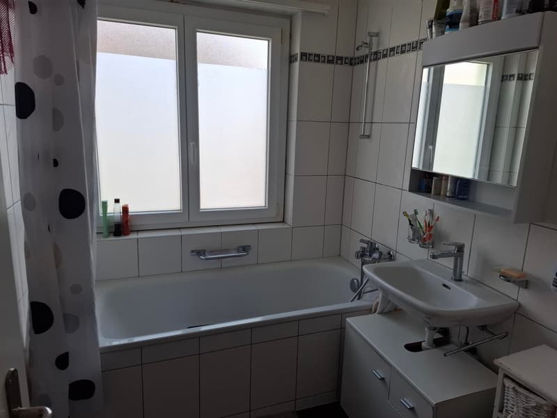 Bathroom: white tiles, bathtub, sink, mirror, window with blinds, toiletries on the window sill