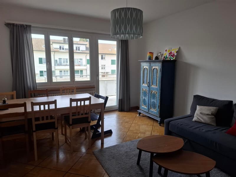 A room with wooden flooring and large windows, dining table with chairs, wooden cabinet, sofa with pillows, and wooden tables.