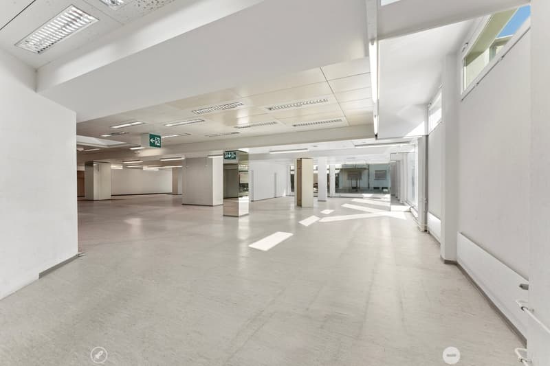 Empty room with white walls, white floor, and white ceiling. There are windows on the right side and exit signs on the ceiling.