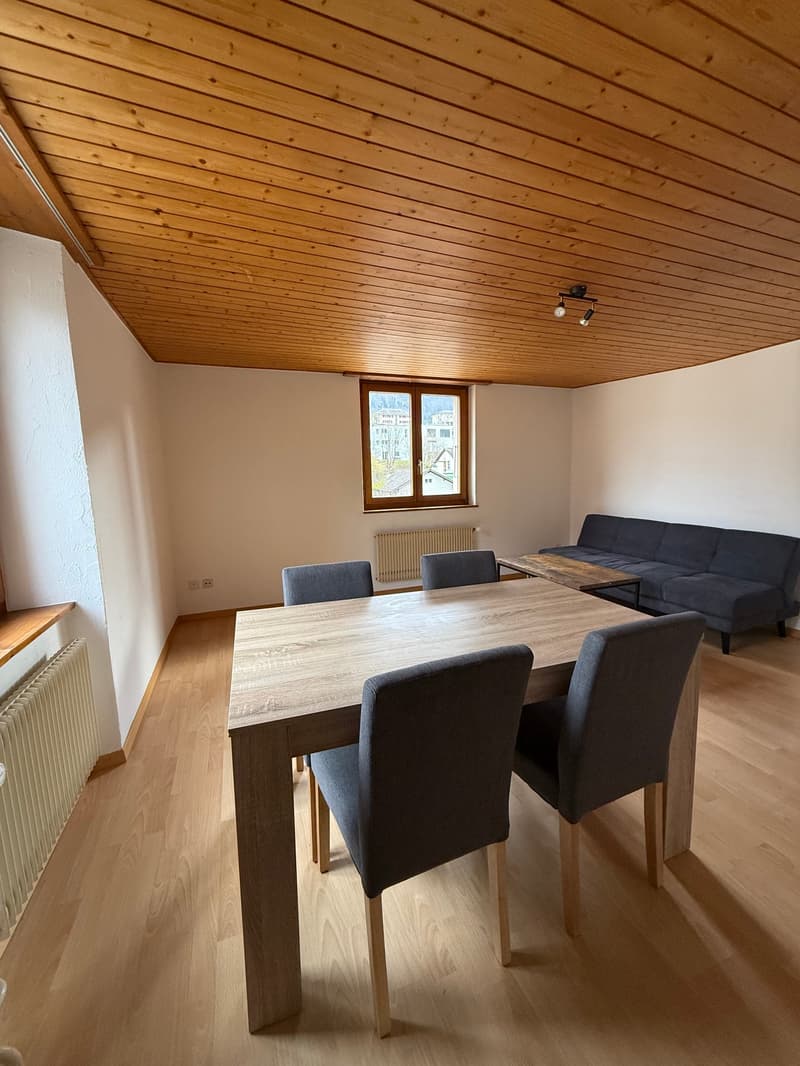 Dining area with a wooden table and chairs, a couch, and a coffee table. Wooden ceiling and white walls. A radiator is on the left wall.