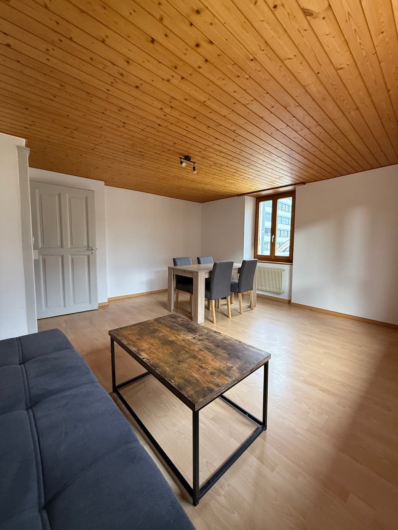 Spacious room with wooden floor, wooden ceiling, a blue couch, a coffee table, a dining table with chairs, a radiator, and a ceiling lamp.