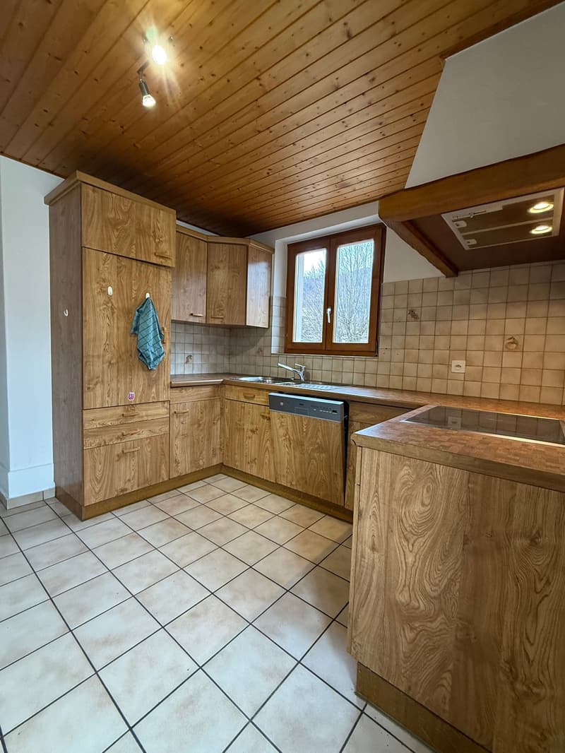 A kitchen with wooden cabinets, tiled floor, and a sink with a faucet. There is a dishwasher under the countertop, and a towel is hanging on a cabinet door. The window provides natural light, and the ceiling has a light fixture.