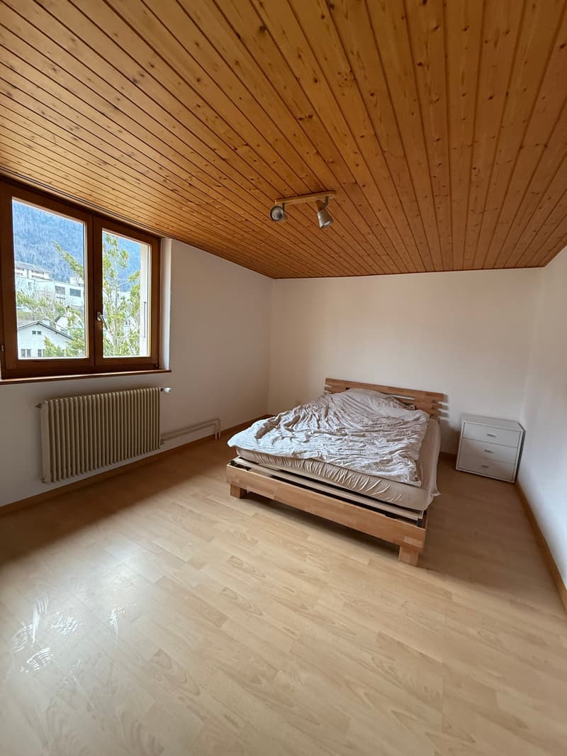 Empty room with wooden floor, white walls, wooden ceiling, a bed, a nightstand, and a radiator.