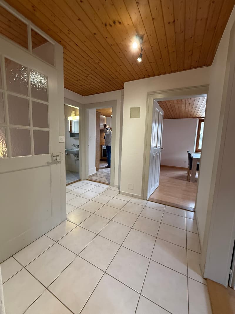 Empty room with white tiled floor, white walls, wooden ceiling, and two doors. One door leads to a bathroom with a sink and mirror, and the other to a kitchen with a sink, faucet, and a person standing. There is also a dining table and chairs in the adjac