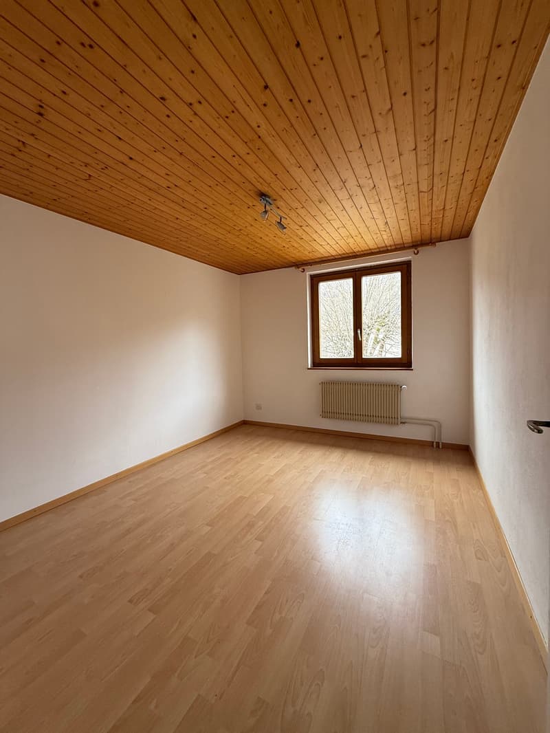 Empty room with wooden floor, white walls, wooden ceiling, and a window with a radiator below.