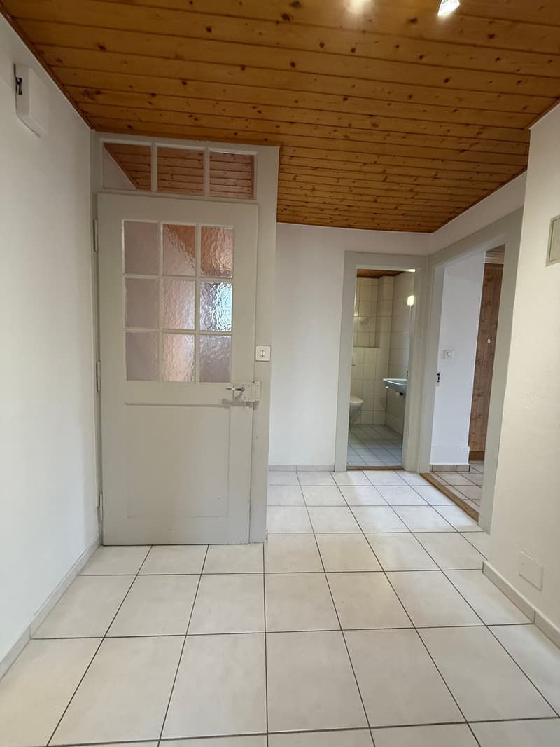 An empty room with white tiled flooring, white walls, and a wooden ceiling. There is a white door with glass panels and a lock. On the right side, there is a small room with a toilet and a sink.