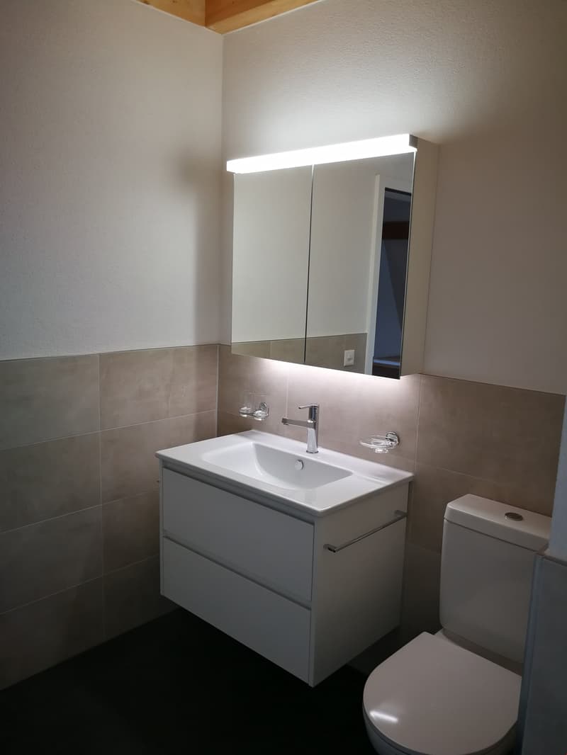 Bathroom with white sink, mirror, and toilet. The sink has a faucet and soap dish. The mirror has a light above it. The walls are tiled.