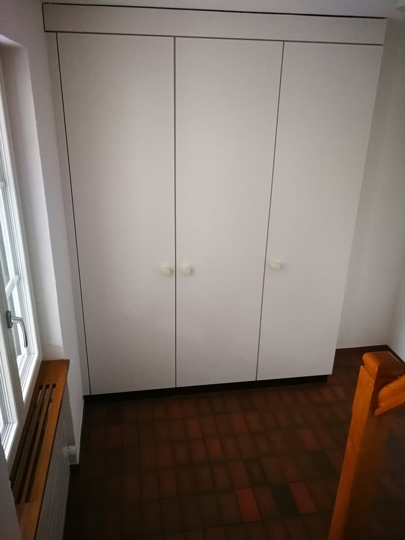 An empty room with white built-in wardrobes, a window, and a wooden bench. The floor is tiled.