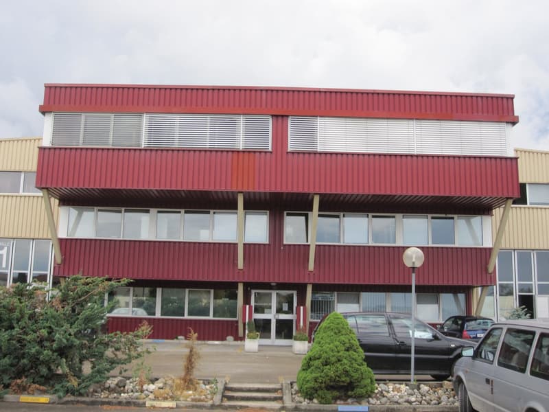 A multi-story commercial building with a red metal exterior and large windows. The building has a main entrance with steps leading up to it. There are some landscaping elements like shrubs and rocks around the building. Several vehicles are parked in the