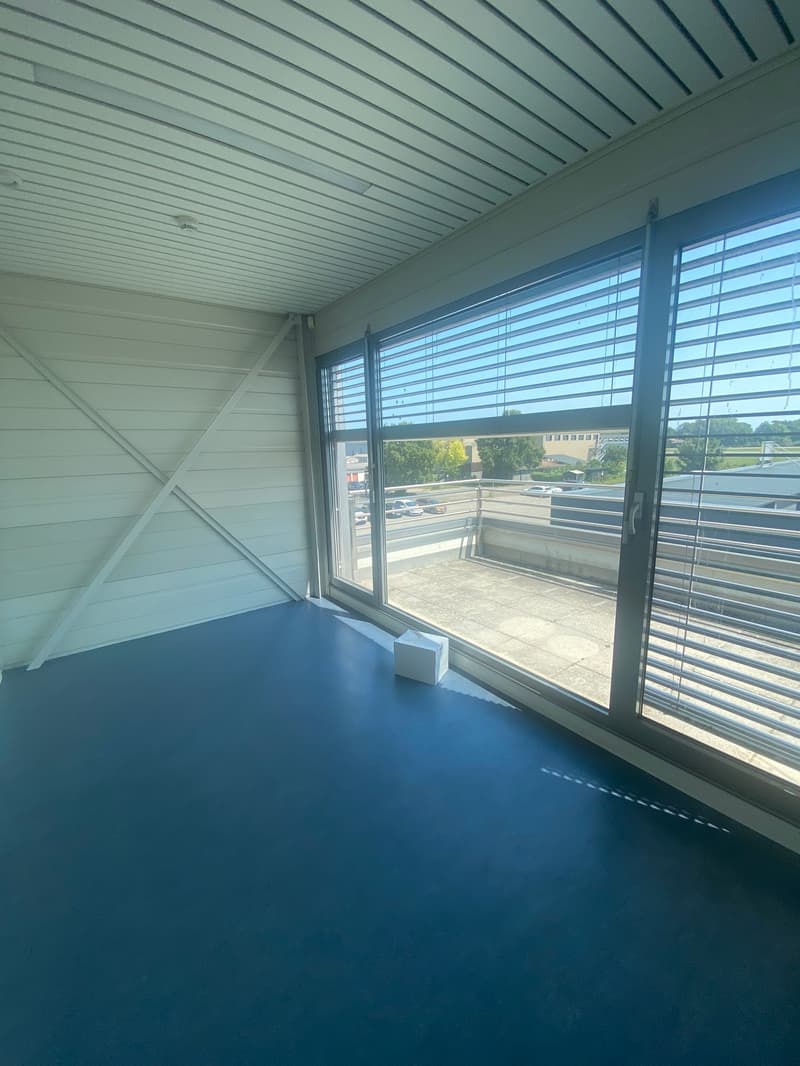 An empty room with blue floor and white wall. There is a large window with blinds and a tissue box beside it. The room has a ceiling with white beams and an exit. Outside the window, there is a building, parking lot, and cars. A building with a gray roof