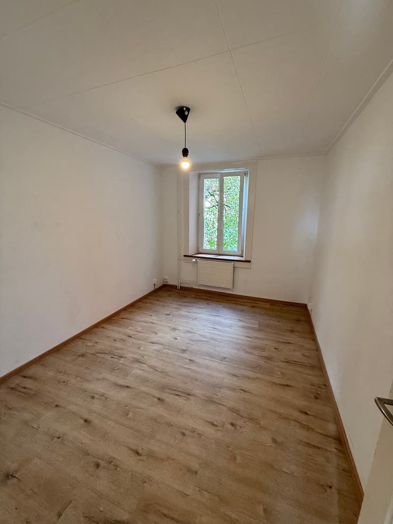 Empty room, wooden floor, white walls, hanging light, window, radiator.
