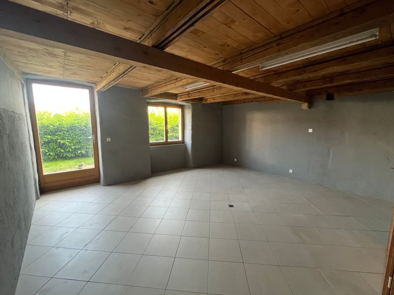 This image shows a large, open room with a wooden ceiling and exposed beams. The room has gray walls and a tiled floor. There are two large windows that provide natural light and views of the surrounding greenery outside. The room appears to be in a state