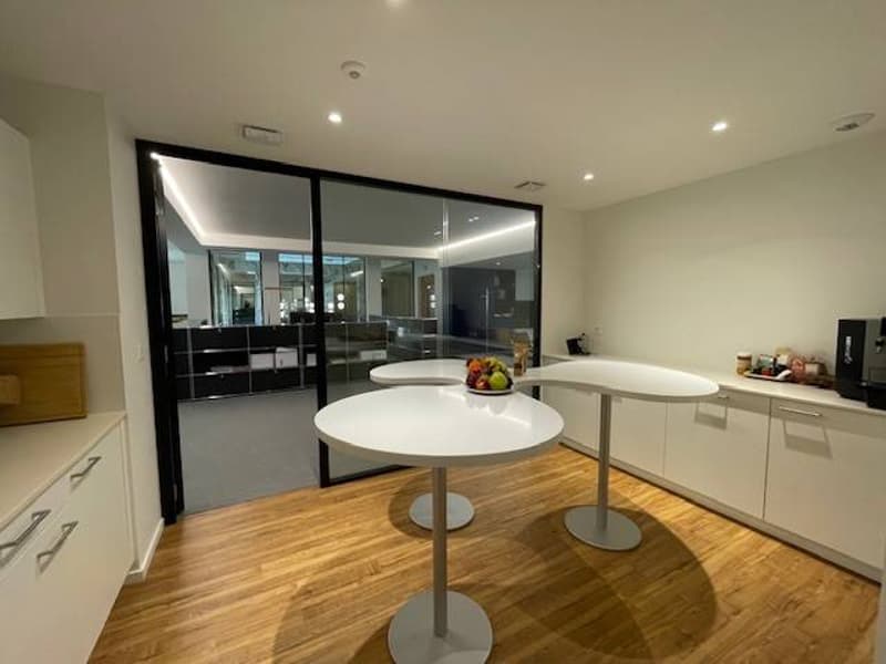 modern kitchen with white cupboards, two white tables, coffee machine, bowl of fruit, wooden floor