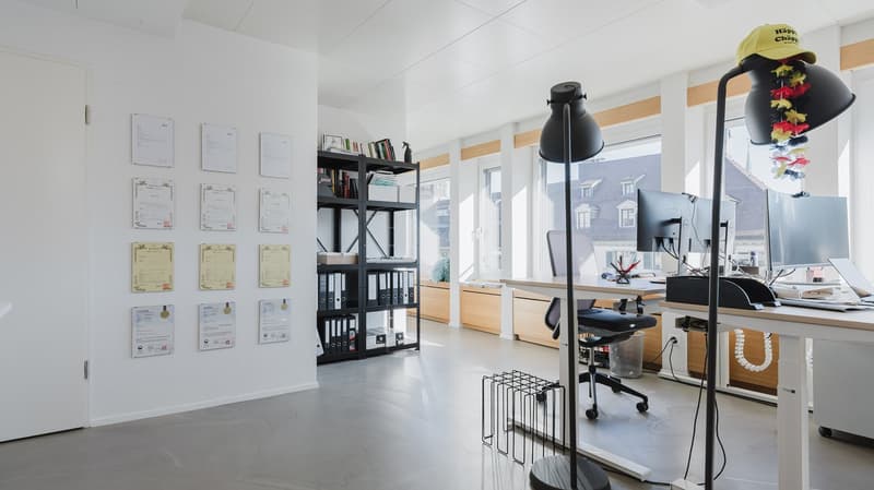 modern office room with white walls, gray floor, large windows, desk, office chair, computer, lamp, shelf with books and files, bulletin board with papers