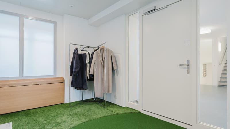 Empty room with clothes rack, green carpet, white walls, large windows, wooden bench, glass door