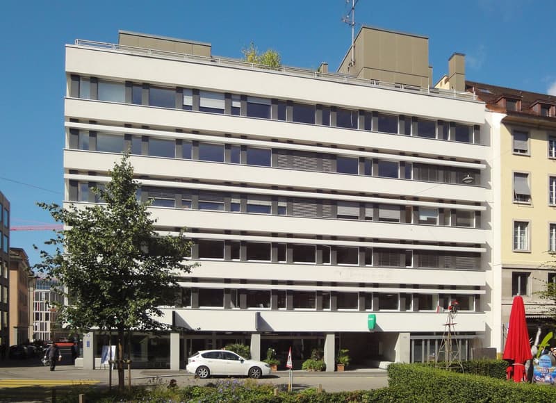 Modern white building with glass windows, rooftop terrace, plants in front, parked car