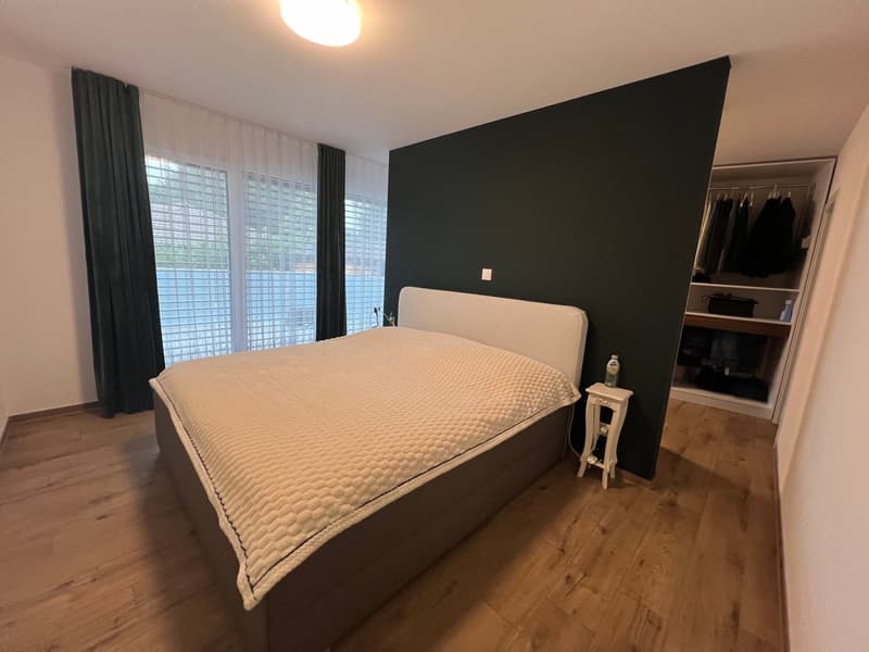 large bed, white headboard, beige bedspread, bedside table, open wardrobe with clothes