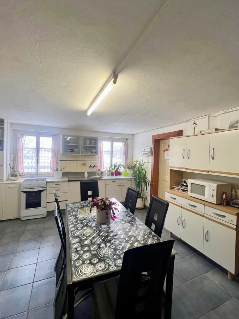 A modern kitchen with a large table and dining chairs, white cabinets, a gas stove, and a sink. There is a microwave on top of the lower cabinets, a potted plant in the corner, and windows with pink curtains.
