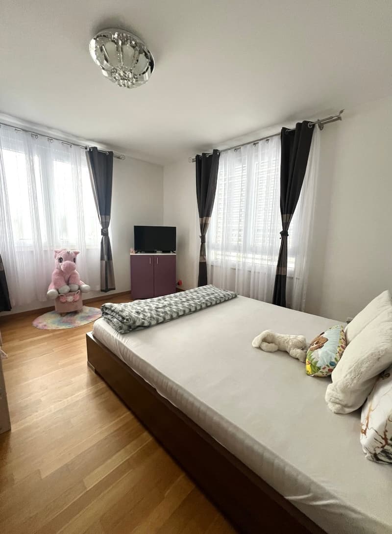 Bedroom with wooden floor, white walls, bed, blanket, pillows, stuffed toys, cabinet, TV, and curtains.