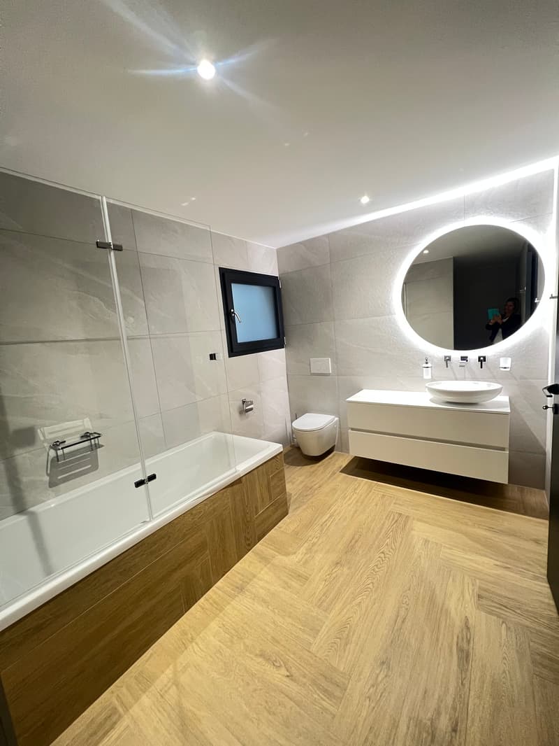 modern bathroom with white bathtub, white sink, white toilet, large mirror, wooden floor