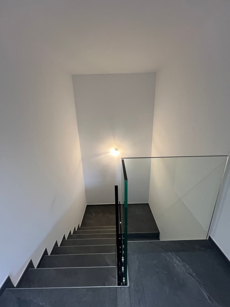 Modern staircase with glass railing, black marble steps, white walls, and a ceiling light