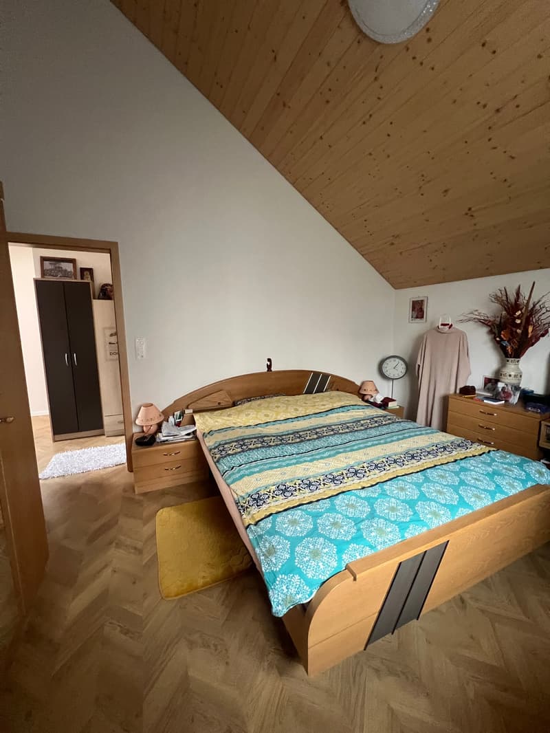 Bedroom with wooden bed, wooden nightstands, wooden wardrobe, wooden floor, white walls, wooden ceiling, lamp, clock, flower vase, picture frame, white rug, and a door.