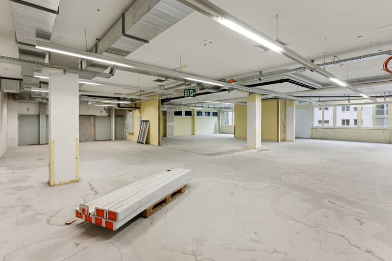 Empty room with concrete floor, metal beams, and fluorescent lights on the ceiling, a stack of white materials on a pallet in the center.