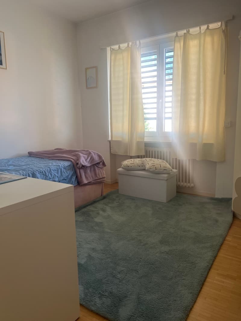 Spacious bedroom with white walls, large windows, hardwood floor, queen-size bed with purple bedding, and a storage box at the foot of the bed.