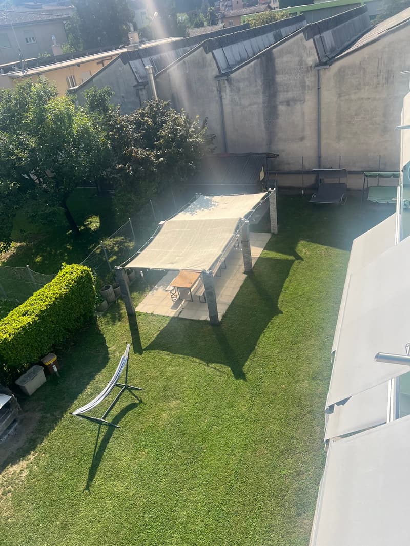 Outdoor patio or courtyard with a grassy lawn, a covered seating area, and some parking spaces. The space is surrounded by buildings and has a view of the trees and greenery outside.