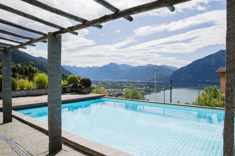 Outdoor pool with a view of the mountains and valley below. The pool is surrounded by a wooden pergola structure and has a tiled deck area. There are potted plants and landscaping around the pool area.