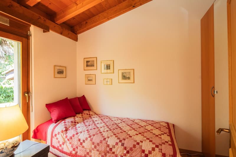 Cozy bedroom with wooden ceiling beams, white walls, framed artwork, red bedding, and a window providing natural light