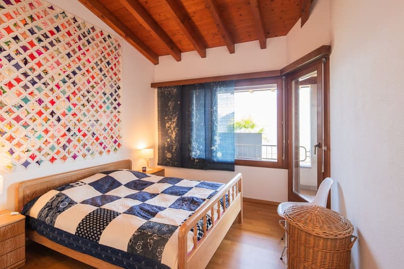 Spacious bedroom with wooden beamed ceiling, large window with balcony access, patchwork quilt on the bed, and a wicker basket in the corner.