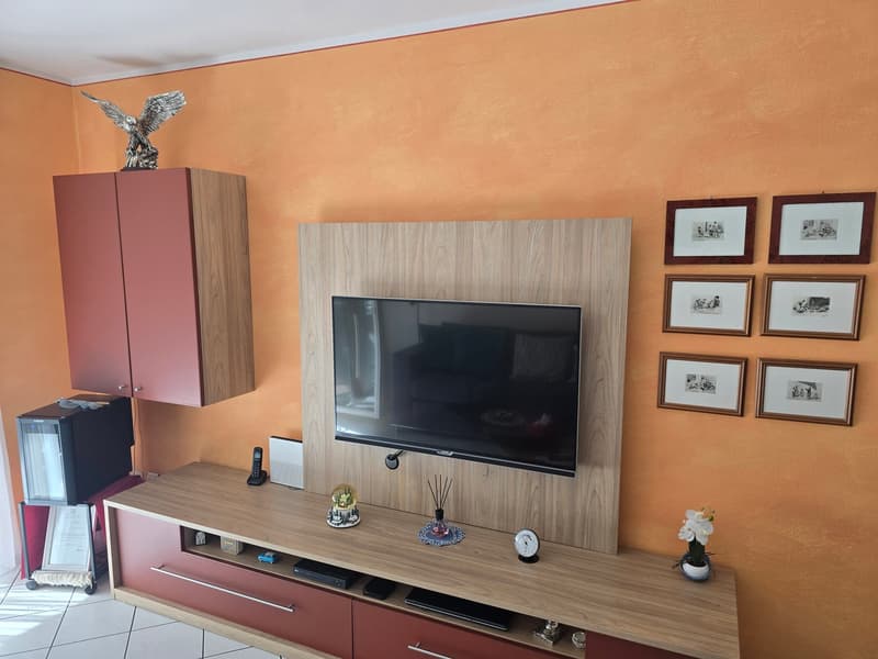 Living room with orange walls, wooden entertainment center, flat screen TV, wooden cabinets, framed pictures, decorative items, and a flower vase.