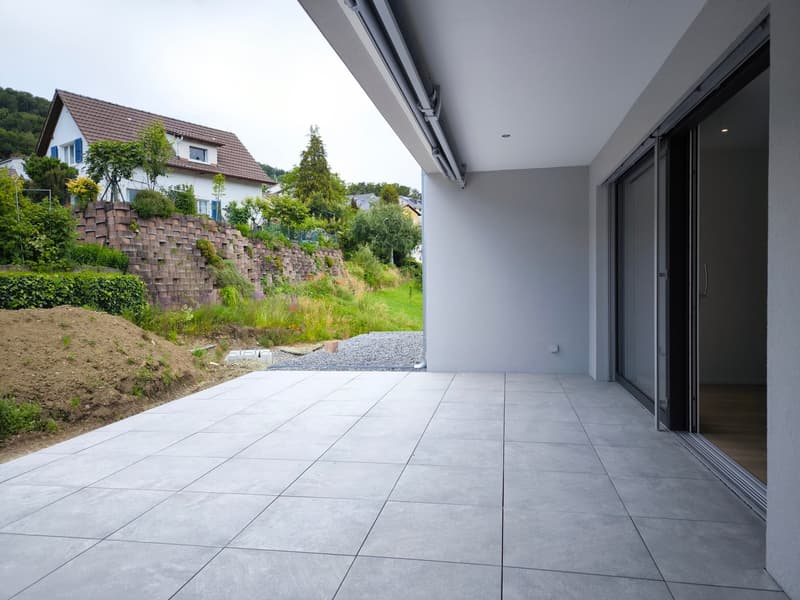 An empty terrace with gray tiles, a glass door, and a view of a house and garden.