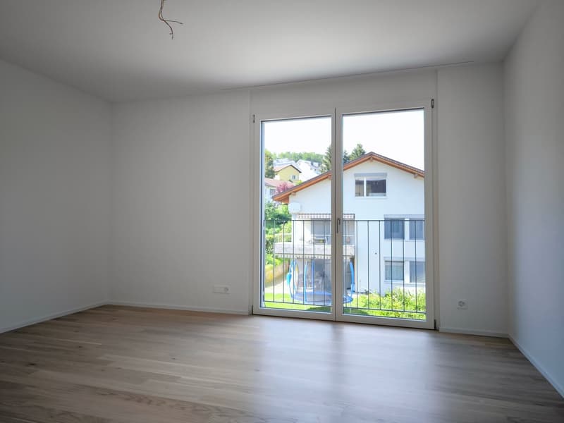 Empty room with white walls, wooden floor, and sliding glass doors leading to a balcony with a view of a house and garden.