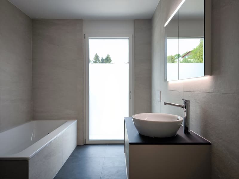 Modern bathroom with a bathtub, a sink, a faucet, and a mirror. The walls and floor are tiled in a light gray color.