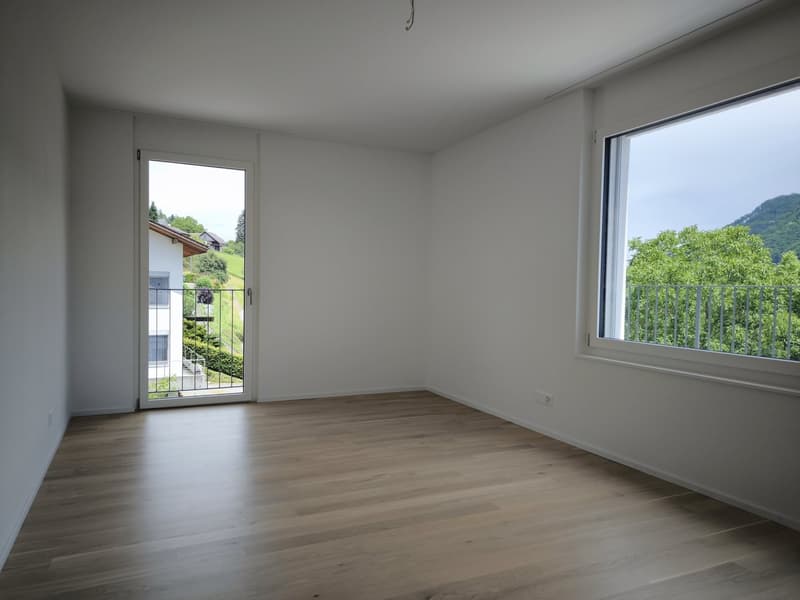 Empty room with white walls, wooden floor, sliding glass door leading to a balcony, and a large window with a view of the outdoors.